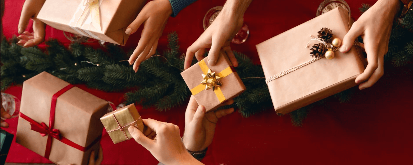 several pairs of hands are sharing brown-paper wrapped gifts over a red and green holiday table setting