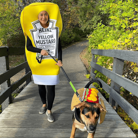 A female dressed in yellow mustard onesie Halloween costume. Her dog is dressed in a rainbow costume. 